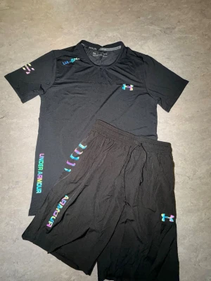 Svart träningsset från Under Armour - Säljer ett svart träningsset från Under Armour med t-shirt och shorts. Setet har färgglada loggor och textdetaljer i neonfärger. Materialet är lätt och snabbtorkande, perfekt för träning eller löpning. T-shirten har rund hals och shortsen har resår i midjan.