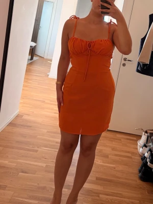 Orange kort klänning med knytband - Snygg orange kort klänning med smala axelband och knytband både framtill och på axlarna. Klänningen har en figurnära passform och är perfekt för varma dagar. Tillverkad i ett lätt material som känns skönt mot huden.Se bilder för mindre defekter.