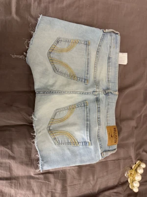 Ljusblå jeansshorts från Hollister - Säljer ett par ljusblå jeansshorts från Hollister med råa, fransiga kanter och klassiska fem fickor. Shortsen har bälteshällor, knapp och dragkedja framtill samt snygga slitningar för en avslappnad look.
