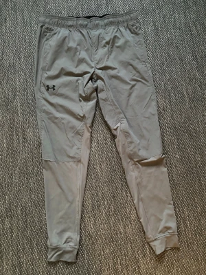 Grå träningsbyxor från Under Armour - Snygga grå träningsbyxor från Under Armour med smal passform och resår i midjan. Byxorna har muddar vid bensluten och är tillverkade i ett lätt och stretchigt material som passar perfekt för gymmet eller löprundan. Diskret logga på vänster lår.