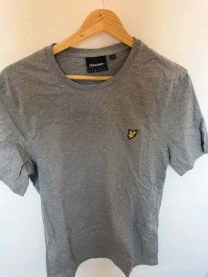 Grå t-shirt från Lyle & Scott - Snygg grå t-shirt från Lyle & Scott med klassisk rund hals och korta ärmar. På bröstet sitter den ikoniska gula loggan med örn. Tillverkad i mjukt bomullsmaterial som känns skönt mot huden. Perfekt basplagg till garderoben.