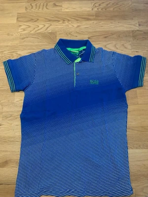 Blå prickig piké från Hugo Boss/ Som S storlek stretchiga  - Snygg blå pikétröja från Hugo Boss med vitt prickigt mönster och gröna detaljer på kragen. Tröjan har klassisk pikékrage, knappar framtill och logga broderad på bröstet. Perfekt för en stilren och sportig look.