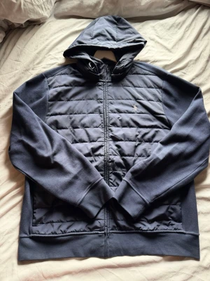 Cardigan från Polo Ralph Lauren - Snygg mörkblå cardigan med huva från Polo Ralph Lauren. Jackan har quiltad front, dragkedja hela vägen och broderad logga på bröstet. Tillverkad i ett mjukt och vadderat material som håller värmen under kalla dagar. Perfekt för kommande höst och vinter. 