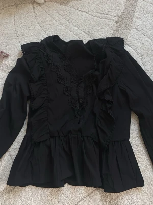 Svart blus med volanger och spets - Snygg svart blus med långa ärmar, volangdetaljer och spets längs ringningen. Blusen har knappar framtill och ett lätt utställt midjeparti som ger en feminin siluett. Perfekt att styla med jeans eller kjol för en trendig look.