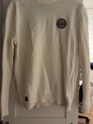 Vit stickad tröja från Morris - Vit stickad tröja från Morris med rund halsringning och ribbade muddar. Tröjan har en snygg patch med Morris-loggan på bröstet och är perfekt för en stilren look. Klassisk och enkel design som passar till mycket.