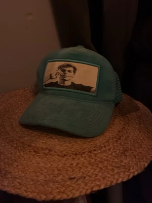 Emilio pendenza keps - Cool turkos truckerkeps med mesh på sidorna och ett svartvitt tryck av en person framtill. Kepsen har böjd skärm och justerbar baksida. Perfekt för dig som vill sticka ut med en unik accessoar. Pris kan diskuteras vid snabb affär. 