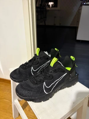 Svarta Nike reacts med neondetaljer - Snygga svarta Nike sneakers med vit broderad swoosh och neonfärgade detaljer på plös och häl. Ovandelen är i mesh för bra andningsförmåga och sulan är chunky och robust. Perfekta för dig som vill sticka ut med coola detaljer och sportig vibe.