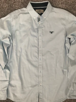 Ljusblå skjorta från Hampton Republic  - Klassisk ljusblå skjorta med button-down krage och broderad logga på bröstet. Skjortan har långa ärmar och knäppning framtill. 