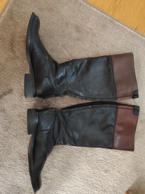 Svarta och bruna ridstövlar i skinn - Snygga höga stövlar i svart och brunt skinn med klassisk ridinspirerad look. Stövlarna har dragkedja på insidan och en liten detalj upptill. Perfekta för dig som gillar stilrena boots med en twist.