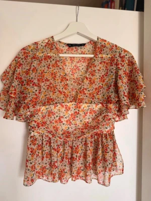 Blommig blus med volangärmar - Söt blommig blus i tunnt, luftigt tyg med volangiga korta ärmar. Mönstret går i orange, rött och gult på ljus botten. Djup v-ringning med snörning framtill och markerad midja ger en feminin siluett. Perfekt för en somrig look.