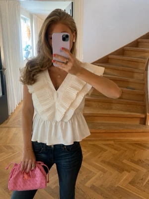 Vit volangblus med v-ringning - Snygg vit blus med breda volangdetaljer och djup v-ringning. Blusen har en luftig passform och är ärmlös, perfekt för en trendig look. Tillverkad i ett lätt material som känns bekvämt mot huden.