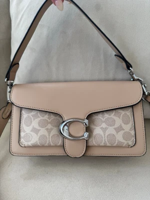 Beige axelväska med silverdetalj - Snygg beige axelväska med bred axelrem och stor silverfärgad C-detalj på locket. Väskan har ett mönstrat tygparti framtill med ljusa toner och släta läderdetaljer. Perfekt för dig som gillar stilrena accessoarer med en twist.