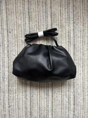 Svart dumpling väska  - Stilren svart kuvertväska i mjukt material med en puffig, veckad design. Väskan har en lång, smal axelrem som kan knytas eller bäras över axeln. Perfekt för dig som gillar minimalistisk och trendig stil.