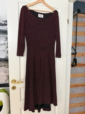 Vinröd glittrig långklänning - Vinröd långklänning med glittrig effekt och trekvartsärm. Klänningen har en rund halsringning och ett lätt utställt fall. Materialet består av nylon, metall och elastan vilket ger en stretchig och skimrande look. Perfekt för dig som vill sticka ut lite extra.