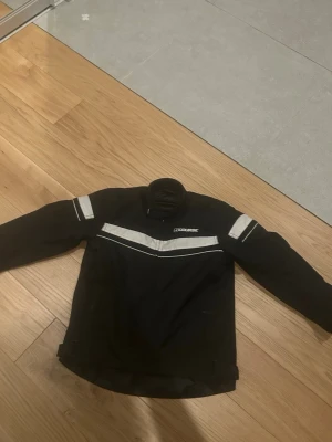 Svart MC-jacka från Course - Snygg svart vindjacka från Course med vita reflexdetaljer på bröstet, ryggen och ärmarna. Jackan har hög krage och raka ärmar, perfekt för blåsiga dagar. Materialet är slitstarkt och jackan har en sportig look.