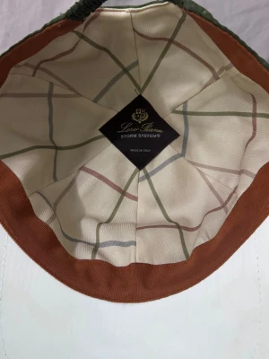 Grön keps från Loro Piana - Snygg grön keps från Loro Piana med Storm System-teknologi. Utsidan är i olivgrönt tyg och insidan har ett rutigt foder i beige, grönt och rött. Skärmen har vit kant och kepsen är tillverkad i Italien. Perfekt accessoar för en stilren look.