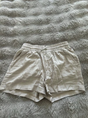 Beige linneshorts med resårmidja - Snygga beige shorts i linne med avslappnad passform och elastisk midja. Shortsen har sidofickor fram och en bakficka, perfekta för varma dagar. Lätta att matcha med olika toppar för en chill sommarstil.