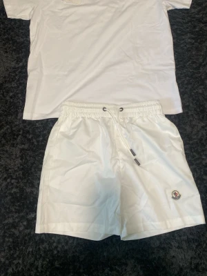 Vita shorts med snörning från Moncler - Säljer ett par fräscha vita shorts från Moncler med elastisk midja och snörning. Shortsen har Moncler-logga på vänster ben och är tillverkade i ett lätt, syntetiskt material som passar perfekt till sommaren.