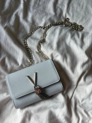 Valentino handväska bag Divina - Snygg ljusgrå axelväska i strukturerat konstläder med silverfärgad kedjerem. Väskan har ett dekorativt V-format metallspänne och hängande kedjedetaljer framtill. Perfekt storlek för det viktigaste och ger en trendig touch till din outfit. Knappt använd och inga defekter, mest suttit som prydnad.