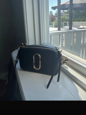 Svart axelväska med gulddetaljer - Snygg svart axelväska i boxig modell med guldfärgad metallogo framtill och dragkedja. Väskan har en bred axelrem med zebramönster och detaljer i guld. Perfekt storlek för det viktigaste och ger en trendig touch till din outfit.
