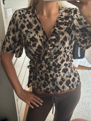 Leopardmönstrad omlottblus - Snygg omlottblus med leopardmönster i beige, brunt och svart. Blusen har korta ärmar, volangdetalj vid ringningen och knyts i midjan för en smickrande passform. Perfekt för dig som vill sticka ut med ett trendigt djurmönster. Storlek 40 men passar mig som har XS i vanliga fall.