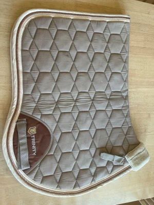 Beige hopp schabrak från Trinity - I storlek full så säljer jag ett beige schabrak från Trinity med geometriskt mönster och ljusbruna detaljer. Schabraket har en mjuk kant och kardborreband för enkel fastsättning. Perfekt till hästutrustningen. Aldrig andvänd 