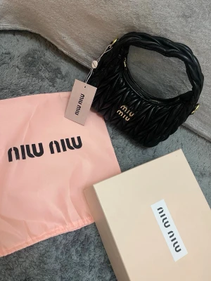Svart quiltad handväska från Miu Miu - Snygg svart handväska från Miu Miu i quiltad design med guldfärgade detaljer och logga framtill. Väskan har en mjuk, vadderad form och kort handtag. Kommer med dustbag och originalkartong. Perfekt accessoar för att lyfta din outfit.