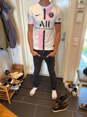 PSG Messi 30 matchtröja vit/rosa - Snygg PSG matchtröja i vitt med rosa och svarta detaljer. Tröjan har tryck med Messi och nummer 30 på ryggen, samt klubbmärke och sponsorloggor framtill. Korta ärmar och sportig passform, perfekt för fotboll eller träning. Trycket i bak är lite utsuddat efter tvätten men fungerar ändå.