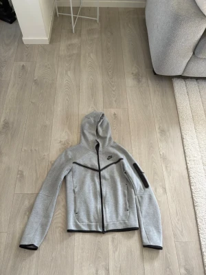 Grå Nike Tech Fleece hoodie - Snygg grå Nike Tech Fleece hoodie med svart dragkedja och detaljer. Hoodien har fickor med dragkedja, svart Nike-logga på bröstet och en extra ficka på ärmen. Tillverkad i mjukt och stretchigt material som passar perfekt till streetwear. Priset kan diskuteras! Levereras inom 24 tim
