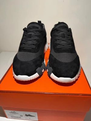 Svarta h sneakers  - Snygga svarta sneakers med ovandel i mesh och mocka, samt en chunky vit sula med svarta detaljer. Skorna har klassisk snörning och en sportig, modern look som passar perfekt till streetwear. Perfekta för dig som gillar stilrena och trendiga sneakers.
