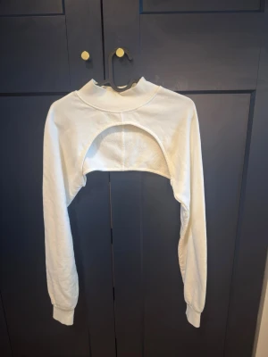 Vit croppad bolerotopp med lång ärm - Trendig vit bolerotopp med croppad passform och långa ärmar. Toppen har en hög ribbad krage och öppen front, perfekt för att styla över linnen eller klänningar. Tillverkad i mjukt bomullsmaterial för en bekväm känsla.