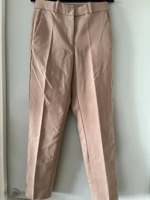 Beige kostymbyxa från H&M - Stilrena beige kostymbyxor från H&M med pressveck och raka ben. Byxorna har hällor för skärp och stängs med hake och dragkedja. Materialet består av bomull och viskosblandning som ger en mjuk och bekväm känsla.