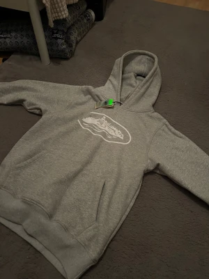 Grå hoodie med tryck och ficka - Säljer en grå hoodie med vit tryckt design på bröstet och stor magficka. Hoodien har en klassisk huva och ribbade muddar vid ärmslut och nederkant. Perfekt för dig som gillar en enkel och stilren look.