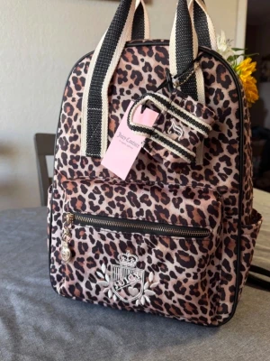 Leopardmönstrad ryggsäck Juicy Couture - Leopardmönstrad ryggsäck från Juicy Couture med svartvita handtag och axelband. Ryggsäcken har en stor ytterficka med dragkedja, Juicy Couture-emblem framtill och matchande liten accessoar. Tillverkad i slitstarkt tygmaterial.