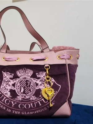 Juicy Couture lila och rosa handväska - Juicy Couture handväska i lila mocka med rosa detaljer och guldiga metalldelar. Väskan har ett stort broderat emblem framtill, rosa handtag och en dekorativ nyckel- och hjärthänge i guld och rosa. Perfekt för dig som gillar statement-accessoarer.