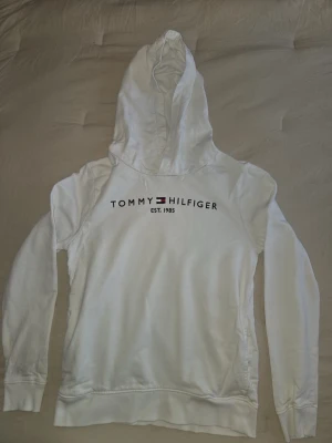 Vit hoodie från Tommy Hilfiger - Snygg vit hoodie från Tommy Hilfiger med tryckt logga och texten 'EST. 1985' på bröstet. Klassisk huva och långa ärmar. Tillverkad i mjukt bomullsmaterial som känns skönt mot huden. Perfekt för en avslappnad och stilren look.