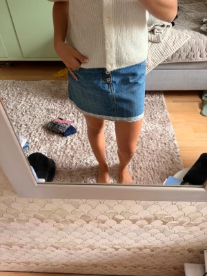 Jeanskjol - Säljer en kort blå jeanskjol med vit spetskant längst ner. Kjolen har knapp- och dragkedjestängning framtill och klassisk femficksdesign. Perfekt för dig som gillar en enkel men söt denimlook.