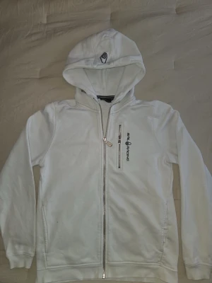 Vit hoodie från Sail Racing - Säljer en vit hoodie från Sail Racing med dragkedja framtill och en liten bröstficka med logga. Hoodien har en broderad detalj på huvan och är tillverkad i mjukt material med fleece på insidan. Perfekt för dig som gillar stilren och sportig look.