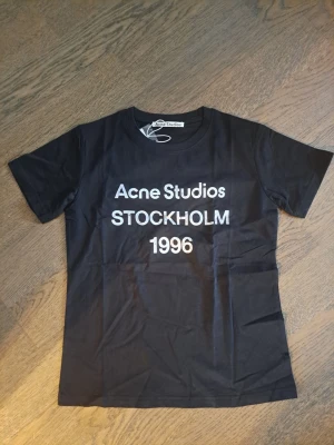 Svart Acne Studios t-shirt - Snygg svart t-shirt från Acne Studios med vit text 'Acne Studios STOCKHOLM 1996' på bröstet. Klassisk passform med rund hals och korta ärmar. Perfekt till jeans eller kjol för en clean och trendig look.