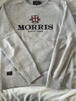 Grå stickad tröja från Morris - Snygg grå stickad tröja från Morris med broderad logga och Union Jack-detalj på bröstet. Tröjan har rund halsringning, ribbade muddar och nederkant. Perfekt för en stilren och avslappnad look. Knappt använd, har legat i garderoben majoriteten av dens livslängd