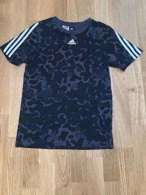 Adidas T-shirt - Snygg svart t-shirt från Adidas med mörkgrått camomönster och klassiska vita ränder på axlarna. Adidas-logga på bröstet. Tillverkad i ett mjukt och lätt material som passar perfekt till en sportig stil. Billig och bra skick!