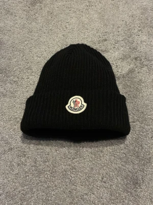 Svart Moncler mössa i ull - Svart ribbstickad mössa från Moncler med klassisk uppvikt kant och broderad logotyp framtill. Tillverkad i mjuk ull som håller dig varm och stilren under kalla dagar. Perfekt accessoar för att lyfta din vinterlook.