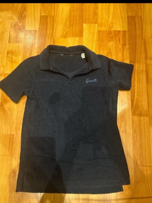 Svart ribbad polotopp från Gant - Den är 134 cm lång och 128 cm lång Snygg svart ribbad polotopp från Gant med korta ärmar och klassisk krage. Toppen har diskret broderad logga på bröstet och är tillverkad i ett mjukt material som ger en bekväm passform. Perfekt till jeans eller kjol för en stilren look.