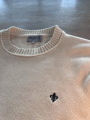 Beige stickad tröja Morris - Klassisk beige stickad tröja från Morris med rund halsringning och ribbade muddar vid ärmslut och nederkant. Tröjan har en liten svart broderad logga på bröstet och är tillverkad i mjukt material som passar perfekt till lager-på-lager.