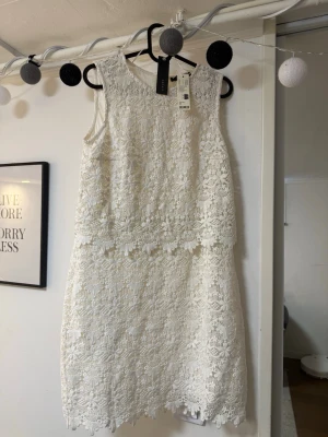 Vit spetsklänning ärmlös - Säljer en elegant vit klänning i spets med ärmlös design och rund hals. Klänningen har ett vackert blommigt spetsmönster och en rak, kort siluett. Perfekt för sommarens alla tillfällen!