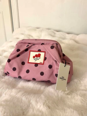 Clutch från Beck söndergaard - Söt rosa clutch med mörklila prickar från Beck Söndergaard. Väskan har en broderad patch med röda blommor på framsidan och ett rymligt fack med dragkedja inuti. Perfekt för skolan. Helt ny med prislapp och nypriset är: 749kr❣️