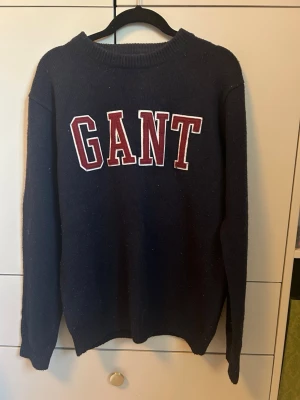 Mörkblå stickad tröja från GANT - Säljer en mörkblå stickad tröja från GANT med stora röda och vita bokstäver på bröstet. Tröjan har rund halsringning, långa ärmar och ribbade muddar vid ärmslut och nederkant. Perfekt för en avslappnad och stilren look.