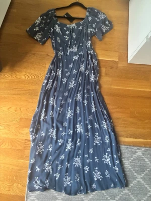 Blå maxiklänning med blommönster - Supersöt blå maxiklänning med vita småblommor och korta puffärmar. Klänningen har en markerad midja och knytband i ryggen för extra passform. Tunt, luftigt tyg som ger ett flowy intryck. Perfekt för sommarens alla tillfällen!