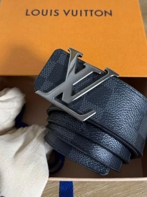 Svart Louis Vuitton bälte - Snyggt svart bälte från Louis Vuitton med klassiskt rutigt mönster och stor LV-logga i silver på spännet. Perfekt accessoar för att lyfta din outfit med en exklusiv touch. Bältet har en bred design och är gjort i skinn.