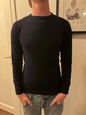 Mörkblå stickad tröja - En stilren mörkblå stickad tröja med rund hals och långa ärmar. Tröjan har en smal passform och är perfekt att matcha med jeans. Materialet är mjukt och bekvämt, troligen bomull eller ullblandning.